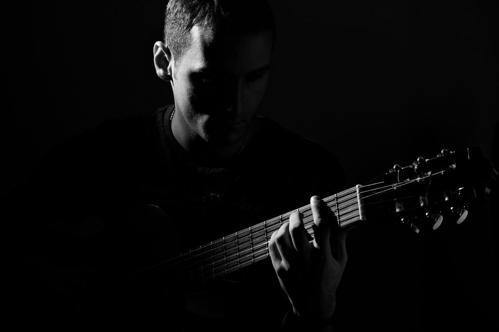 Das Schwarz-Weiß-Foto zeigt eine Person, die eine akustische Gitarre spielt. Die Szene ist in dramatischem, niedrigem Licht gehalten, wodurch starke Schatten entstehen und die Konturen von Gesicht, Hand und Gitarrensaiten eindrucksvoll hervorgehoben werden. Das Gesicht der Person ist teilweise im Schatten verborgen, was dem Bild eine nachdenkliche, fast meditative Stimmung verleiht. Die Finger liegen auf dem Griffbrett – ein Moment des Spielens oder Übens, eingefangen mit künstlerischer Präzision.